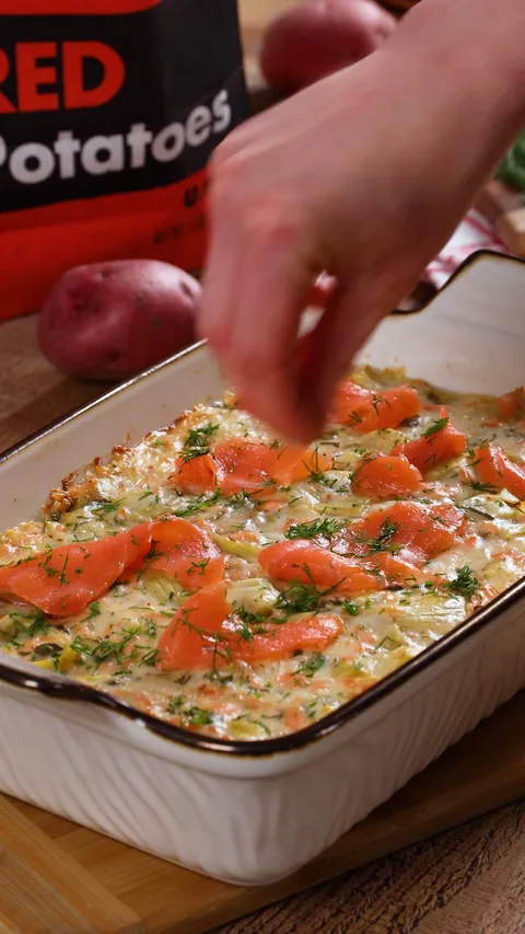 The gratin in its baking dish, topped with smoked salmon and dill, a bag of Spud & Root Red Potatoes behind it.